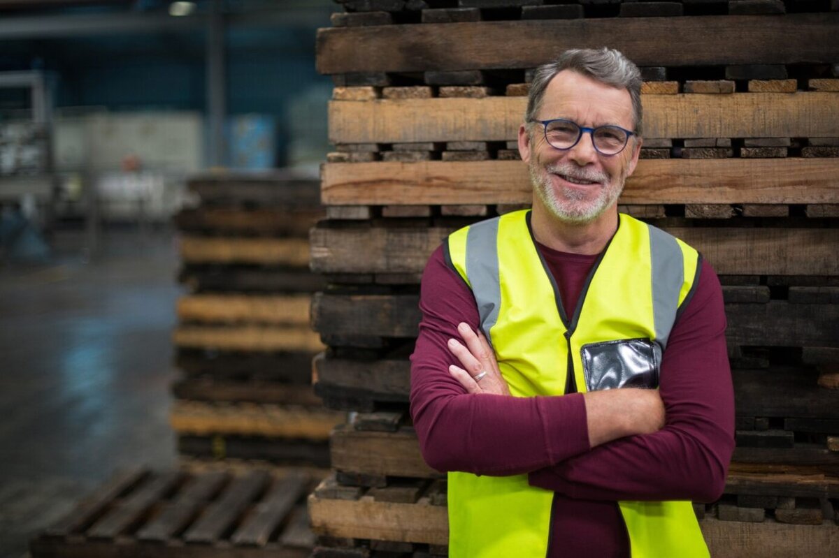 Older man wearing blue glasses and a yellow vest standing in front of wooden planks in a warehouse