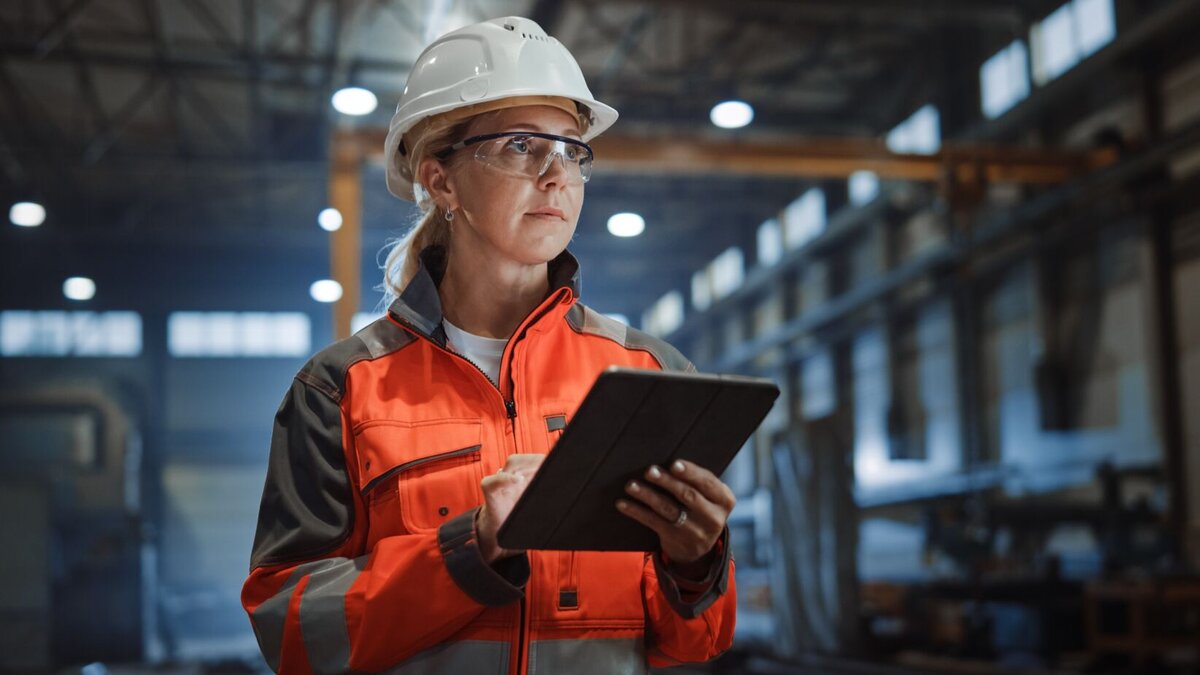 woman in white construction hat and orange jumpsuit holding an ipad in a warehouse