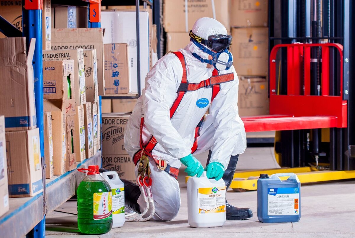 A person in a hazmat suit kneeling by bottles of chemicals in a warehouse
