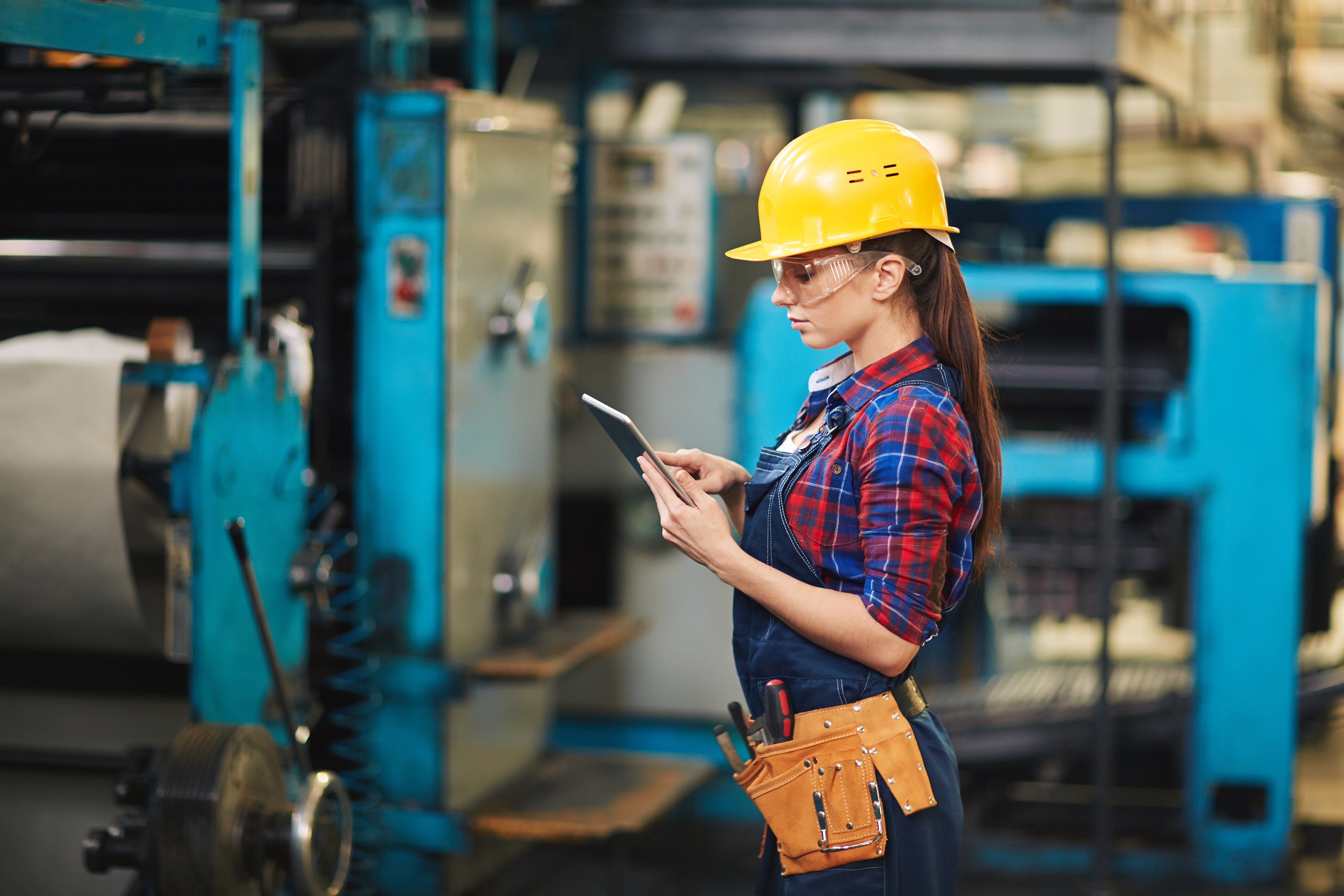 woman in a yellow construction hat wearing a checkered shirt and a toolbelt, looking at an iPad in a manufacturing facility
