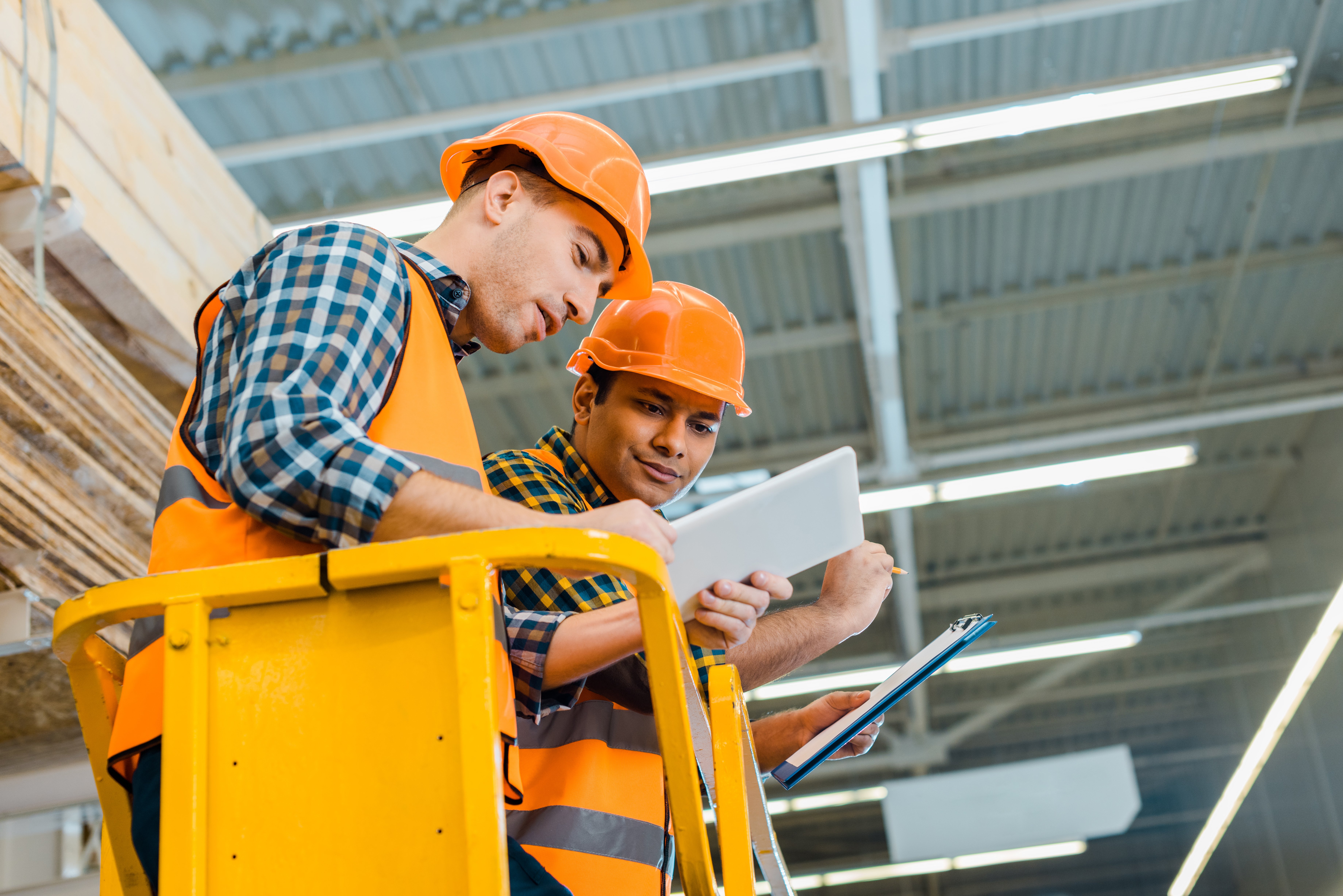 two workers in orange construction hats on a lift in a warehouse looking at an iPad