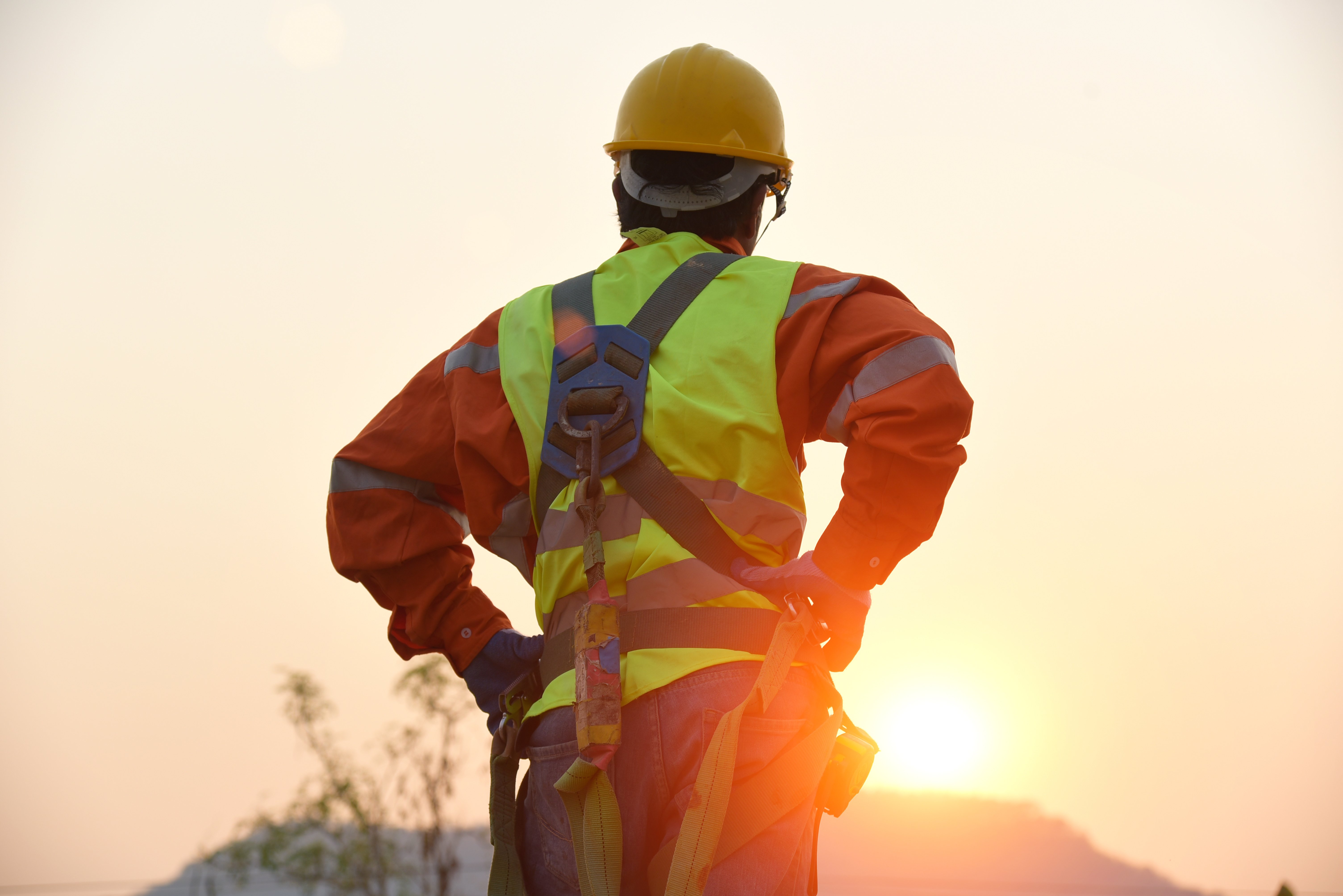 Person wearing a harness, orange jumpsuit, and yellow construction hat looking at the sunset outside