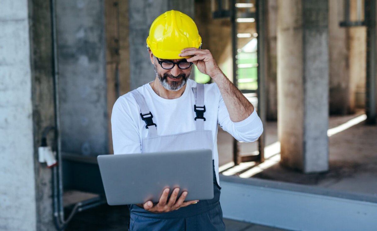 Man with yellow hardhat standing with an open silver laptop in his hands